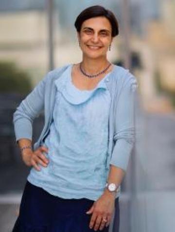 Tulia Falleti is pictured from the waist up, wearing a blue shirt and a cardigan, with right hand on hip and left hand hanging down displaying a wrist watch.